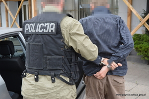 Zdjęcie poglądowe, przedstawiające policjanta w kamizelce z napisem POLICJA. Policjant prowadzi zatrzymanego.