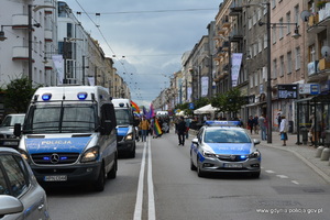 Zdjęcie poglądowe, przedstawiające ulicę Świętojańską. Przez ulicę jadą w tym samym kierunku radiowozy policyjne zajmując oba pasy ruchu. Za radiowozami  idą umundurowani policjanci, którzy zabezpieczają marsz, a także uczestnicy marszu.