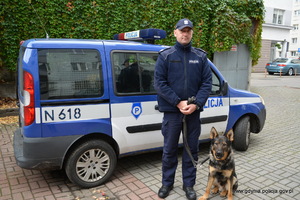 Zdjęcie poglądowe, przedstawiające policjanta z psem służbowym, który podjął trop i dzięki temu znaleziono zaginioną 21-latkę. Policjant z psem stoi na tle oznakowanego radiowozu.