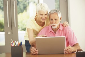 Zdjęcie poglądowe, przedstawiające starszą kobietę i starszego mężczyznę. Mężczyzna siedzi przy biurku przed laptopem, kobieta opiera się o niego i razem patrzą w ekran laptopa.
