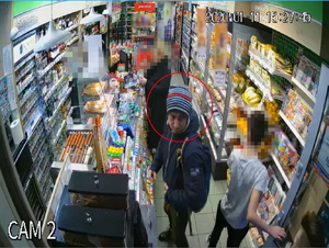 Zdjęcie poglądowe, przedstawiające mężczyznę, który stoi w sklepie tyłem do lodówki, a przodem do półek z alkoholami. Mężczyzna ma na sobie ciemną kurtkę, ciemne spodnie i czapkę. Patrzy w lewą stronę.