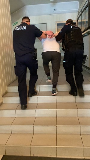 Zdjęcie przedstawiające dwóch policjantów prowadzących po schodach zatrzymanego mężczyznę.