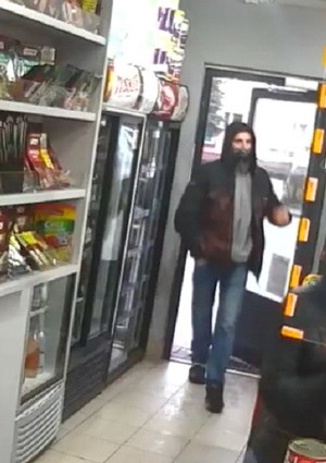 Zdjęcie przedstawiające mężczyznę, który wszedł do sklepu. Mężczyzna ma na sobie ciemne buty, ciemną kurtkę z kapturem na głowie, maseczkę na twarzy oraz niebieskie jeansy.