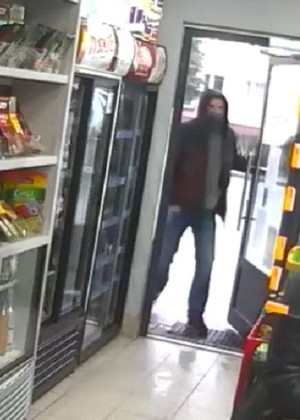Zdjęcie przedstawiające mężczyznę, który wchodzi do sklepu. Mężczyzna ma na sobie ciemne buty, ciemną kurtkę z kapturem na głowie, maseczkę na twarzy oraz niebieskie jeansy.