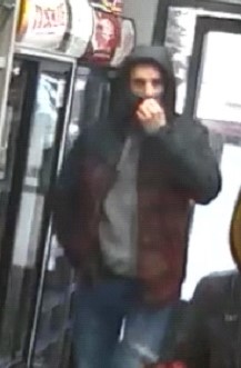 Zdjęcie przedstawiające mężczyznę, który jest w sklepie. Mężczyzna ma na sobie ciemną kurtkę z kapturem na głowie, maseczkę na twarzy oraz niebieskie jeansy.
