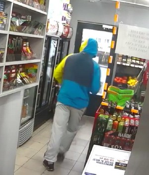 Zdjęcie przedstawiające tył mężczyzny, który idzie w kierunku drzwi wyjściowych ze sklepu. Mężczyzna ma na sobie ciemne, sportowe buty, szare spodnie dresowe i niebieską kurtkę z kapturem. Lewy rękaw kurtki jest koloru żółtego, a górna część kurtki jest granatowa. Mężczyzna ma kaptur na głowie.