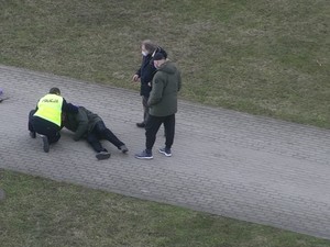 Zdjęcie przedstawiające policjanta udzielającego pomocy mężczyźnie, który leży na chodniku. Sytuacji przypatruje się dwóch przechodniów.