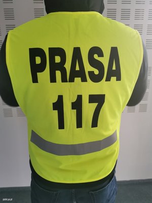 Zdjęcie przedstawiające mężczyznę stojącego tyłem, w kamizelce odblaskowej. Na kamizelce jest napis: PRASA 117