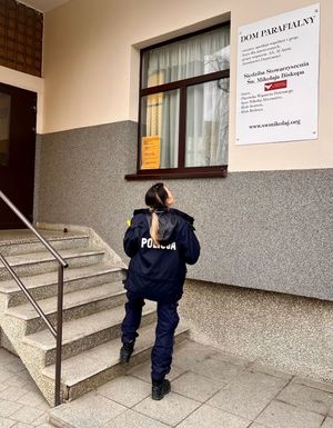 Zdjęcie przedstawiające policjantkę, która stoi prze wejściem do stowarzyszenia i zwrócona jest głową w kierunku tablicy z napisem Dom Parafialny
