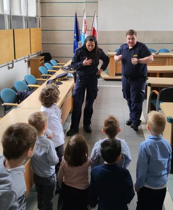 Zdjęcie przedstawiające policjantkę i strażnika miejskiego podczas spotkania z dziećmi. Funkcjonariusze stoją przodem do dzieci i do zdjęcia (policjantka z lewej strony strażnik miejski z prawej strony), natomiast dzieci stoją tyłem do fotografii.