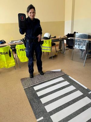 Zdjęcie przedstawiające policjantkę, która stoi przy macie z przejściem dla pieszych i trzyma w rękach znak sygnalizator świetlny dla dzieci