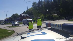 Na zdjęciu widać policjantkę i policjanta stojących na tle radiowozu policyjnego. Policjant sprawdza prędkość nadjeżdżających samochodów urządzeniem do pomiaru prędkości.