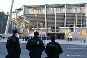 Zdjęcie poglądowe, przedstawiające z tyłu trzech policjantów stojących przodem do stadionu. W tle grupa osób.