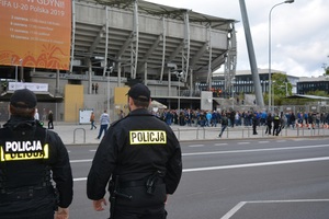 Zdjęcie poglądowe, przedstawiające z tyłu dwóch policjantów stojących twarzą do stadionu. W tle widać kibiców.