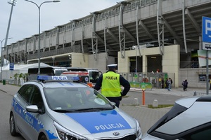 Zdjęcie poglądowe, przedstawiające policjantów na tle radiowozu i stadionu. W tle kibice.