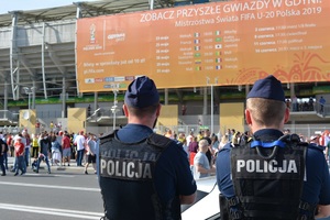 Zdjęcie poglądowe, przedstawiające dwóch policjantów stojących przed stadionem. W tle widać grupę osób.