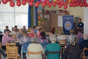 Zdjęcie przedstawia seniorów siedzących przy ławkach ustawionych w tzw. podkowę oraz stojącego policjanta, który prowadzi z prelekcję na temat bezpieczeństwa seniorów.