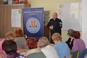 Zdjęcie przedstawia policjanta, który opowiada seniorom o bezpieczeństwie w ruchu drogowym. Przy ławkach ustawionych w kształcie podkowy siedzą osoby. W tle baner Komendy Miejskiej Policji w Gdyni.