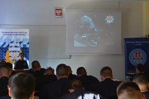 Zdjęcie przedstawia salę lekcyjną. W sali po prawej stronie stoi baner Komendy Miejskiej Policji w Gdyni, po lewej baner Akademii Marynarki Wojennej w Gdyni. Na fotografii można zobaczyć policjantkę siedzącą przy biurku, która omawia prezentację wyświetlaną na tablicy multimedialnej. Przodem do niej, w ławkach  siedzą słuchacze.