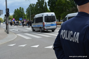 Zdjęcie poglądowe, przedstawiające stojącego policjanta i radiowozy jadące ulicą. W tle przemarsz ludzi.