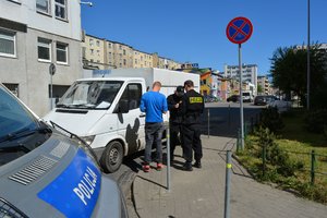 Zdjęcie poglądowe, przedstawiające dwóch policjantów legitymujących mężczyznę. Na zdjęciu widać oznakowany radiowóz, samochodów dostawczych i osiedle bloków mieszkalnych.