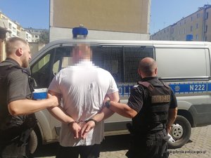 Zdjęcie poglądowe, przedstawiające dwóch umundurowanych policjantów prowadzących do radiowozu mężczyznę, który został zatrzymany za groźby karalne i uszkodzenie samochodu.