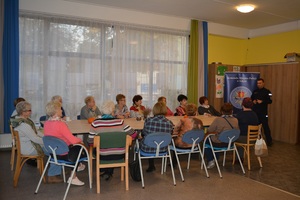 Zdjęcie poglądowe, przedstawiające policjanta, który rozmawia z seniorami. Seniorzy siedzą przy stołach.