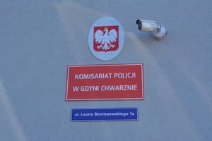 Zdjęcie poglądowe, przedstawiające ścianę budynku komisariatu policji. Na górnej części zdjęcia widać Godło Polski, po prawej stronie od godła widać kamerę. Pod godłem można zobaczyć czerwoną tablicę z białym napisem: Komisariat Policji w Gdyni Chwarznie, a zaraz pod tą tablicą jest niebieska tablica z białym napisem: ul. Leona Staniszewskiego 1A.