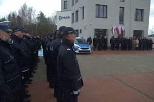 Zdjęcie poglądowe, przedstawiające funkcjonariuszy ustawionych w dwuszeregu przed budynkiem Komisariatu Policji w Gdyni Chwarznie podczas uroczystego otwarcia nowej placówki policji. Na zdjęciu można zobaczyć też oznakowany radiowóz i zaproszonych gości.