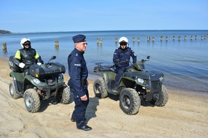 Zdjęcie przedstawiające dwóch policjantów będących na quadach. Obok nich stoi policjant, który patroluje plażę.