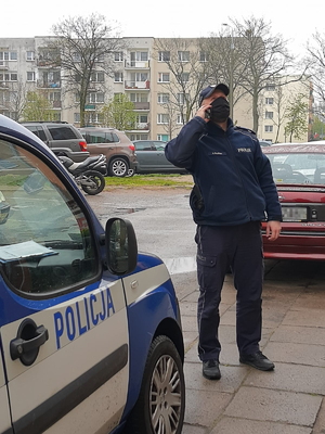 Zdjęcie przedstawiające umundurowanego policjanta na tle radiowozu i zaparkowanych samochodów. Funkcjonariusz ma maseczkę na twarzy i rozmawia przez telefon z osobą będącą na kwarantannie domowej. Policjant patrzy do góry aby zobaczyć swojego rozmówcę.