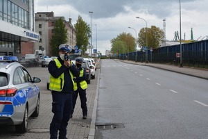 Zdjęcie przedstawiające policjantkę i policjanta ruchu drogowego na tle oznakowanego radiowozu. Policjantka stoi za policjantem. Funkcjonariusz mierzy prędkość nadjeżdżającym samochodom.