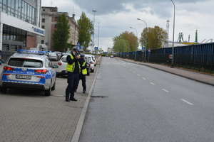 Zdjęcie przedstawiające policjantkę i policjanta ruchu drogowego na tle oznakowanego radiowozu. Policjantka stoi za policjantem. Funkcjonariusz mierzy prędkość nadjeżdżającym samochodom.