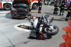 Zdjęcie przedstawiające leżący na ulicy motocykl i samochód osobowy z podniesioną pokrywą silnika oraz pękniętym przednim zderzakiem. W tle widać strażaków oraz wóz Straży Pożarnej i karetkę.