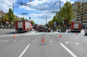 Zdjęcie przedstawiające skrzyżowanie, na którym doszło do wypadku. Na ulicy leży motocykl, obok samochód osobowy z podniesioną pokrywą silnika. Na zdjęciu widać strażaków oraz dwa samochody strażackie i karetkę.