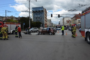 Zdjęcie przedstawiające skrzyżowanie, na którym doszło do zdarzenia drogowego. Na zdjęciu widać policjanta drogówki i strażaków. Ponadto można zobaczyć samochód osobowy, leżący motocykl i dwa wozy strażackie.