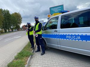 Zdjęcie poglądowe, przedstawiające dwóch policjantów na tle oznakowanego radiowozu. Funkcjonariusze trzymają w rękach tarcze do zatrzymania pojazdu. Jeden z nich mierzy prędkość nadjeżdżających w ich kierunku samochodów.