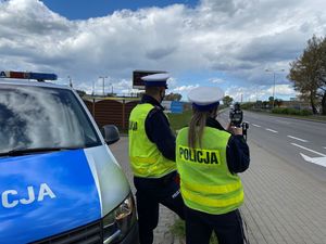 Zdjęcie przedstawiające policjantkę i policjanta. Policjanci stoją tyłem na tle oznakowanego radiowozu. Policjantka mierzy prędkość pojazdów.
