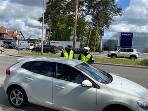 Zdjęcie przedstawiające funkcjonariuszy ruchu drogowego podczas kontroli drogowej kierującego białym samochodem osobowym. Policjanci legitymują kierowcę.