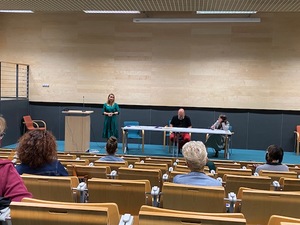 Zdjęcie przedstawiające salę konferencyjną. Na podium jest stół, a przy nim mężczyzna i kobieta. Obok nich stoi kobieta, która prowadzi spotkanie. Przed podium widać publiczność, która bierze udział w posiedzeniu.