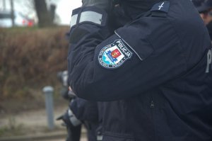 Zdjęcie przedstawiające lewy bok umundurowanego policjanta. Widać zgiętą rękę, a na rękawie naszywkę policyjną.