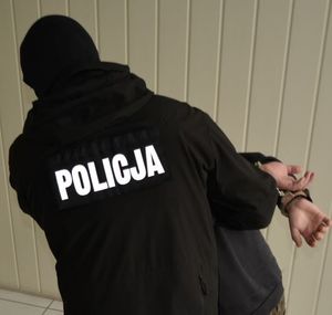 Zdjęcie przedstawiające umundurowanego policjanta. Na zdjęciu widać ręce mężczyzny, którego prowadzi funkcjonariusz.