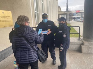 Zdjęcie przedstawiające policjantkę i strażnika miejskiego w trakcie działań. Funkcjonariusze przekazują kobiecie maseczkę.