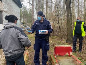 Zdjęcie przedstawiające policjanta, przekazującego osobie bezdomnej maseczkę. Na zdjęciu widać również strażnika miejskiego.