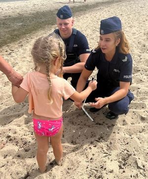 Zdjęcie przedstawiające policjantów na plaży, przekazujących dziecku ulotkę