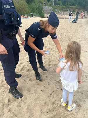 Zdjęcie przedstawiające policjantów na plaży, przekazujących dziecku ulotkę