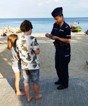 Zdjęcie przedstawiające policjantkę, która przekazuje dzieciom na plaży ulotkę z informacją o konkursie &amp;quot;Artystyczny przeWODNIK&amp;quot;