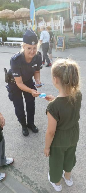 Zdjęcie przedstawiające policjantkę, która przekazuje dziecku na plaży ulotkę z informacją o konkursie &quot;Artystyczny przeWODNIK&quot;