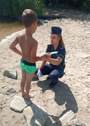 Zdjęcie przedstawiające umundurowaną policjantkę na plaży, która daje dziecku ulotkę z informacją o konkursie