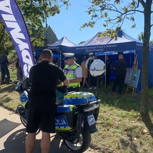 Zdjęcie przedstawiające policjantów podczas wydarzenia pn. Wietrzenie Schronu.. Na zdjęciu widać policjanta i mężczyznę sadzającego dziecko na policyjny motocykl, w tle stoisko policyjne z policjantami i uczestnikami wydarzenia.